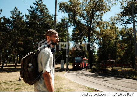 Young man with a backpack standing near tram tracks in a park on a sunny day. Concept of travel, transport, and urban lifestyle Young man with a backpack standing near tram tracks in a park on a sunny day. Concept of travel, transport, and urban lifestyle 141695937