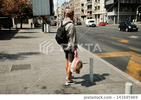 Young man standing on a sunny city street holding a bag of groceries. Concept of urban living, simplicity, and sustainable lifestyle Young man standing on a sunny city street holding a bag of groceries. Concept of urban living, simplicity, and sustainable lifestyle 141695969