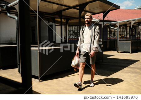 Young man walking through an empty outdoor market holding a plastic bag with groceries. Concept of urban lifestyle, daily errands, and minimalism Young man walking through an empty outdoor market holding a plastic bag with groceries. Concept of urban lifestyle, daily errands, and minimalism 141695970