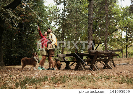 Smiling couple standing in a forest, woman pointing towards trees while two dogs stay nearby. Concept of adventure, nature exploration, and travel Smiling couple standing in a forest, woman pointing towards trees while two dogs stay nearby. Concept of adventure, nature exploration, and travel 141695999