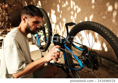 Focused man maintaining his blue mountain bike by spraying lubricant on the chain outdoors. Concept of care, maintenance, and active living Focused man maintaining his blue mountain bike by spraying lubricant on the chain outdoors. Concept of care, maintenance, and active living 141696019