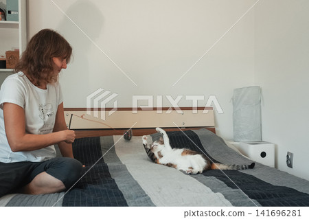 Woman entertaining a playful tabby cat on a bed with an interactive wand toy. Cozy indoor lifestyle scene highlighting enrichment, exercise, bonding, and pet wellbeing Woman entertaining a playful tabby cat on a bed with an interactive wand toy. Cozy indoor lifestyle scene highlighting enrichment, exercise, bonding, and pet wellbeing 141696281