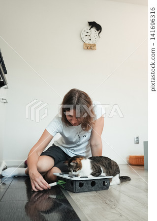 Woman encouraging a tabby cat to explore an interactive puzzle toy indoors. Enrichment activity promotes play, curiosity, and mental stimulation Woman encouraging a tabby cat to explore an interactive puzzle toy indoors. Enrichment activity promotes play, curiosity, and mental stimulation 141696316