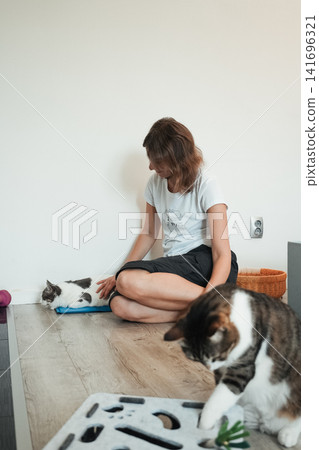Woman relaxing with two domestic cats during indoor playtime at home. Cozy everyday moment showing companionship and pet enrichment Woman relaxing with two domestic cats during indoor playtime at home. Cozy everyday moment showing companionship and pet enrichment 141696321