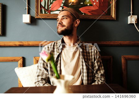 Young man sitting in cozy cafe near wall art, relaxing and thinking. Concept of leisure, lifestyle, and calm moment 141696360