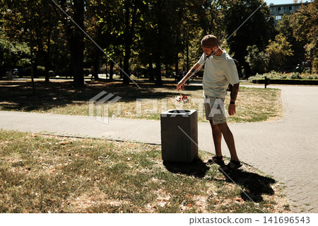 Young man standing in a sunny park throwing garbage into a trash bin. Concept of cleanliness, ecology, and responsibility Young man standing in a sunny park throwing garbage into a trash bin. Concept of cleanliness, ecology, and responsibility 141696543