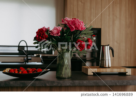 Stylish bouquet of pink and red peonies in a glass vase on modern kitchen counter with strawberries and moka coffee pot. Scandinavian home interior. Stylish bouquet of pink and red peonies in a glass vase on modern kitchen counter with strawberries and moka coffee pot. Scandinavian home interior. 141696889