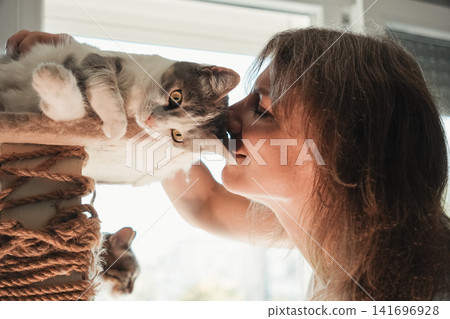 Woman sharing an affectionate moment by kissing her domestic cat resting on a cat tree. Warm indoor lifestyle scene highlighting love, companionship, trust, and pet care Woman sharing an affectionate moment by kissing her domestic cat resting on a cat tree. Warm indoor lifestyle scene highlighting love, companionship, trust, and pet care 141696928