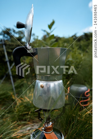 A classic aluminum moka pot geyser coffee maker with an open lid stands on a portable gas burner stove outdoors in the green grass during a mountain hike. A classic aluminum moka pot geyser coffee maker with an open lid stands on a portable gas burner stove outdoors in the green grass during a mountain hike. 141698465