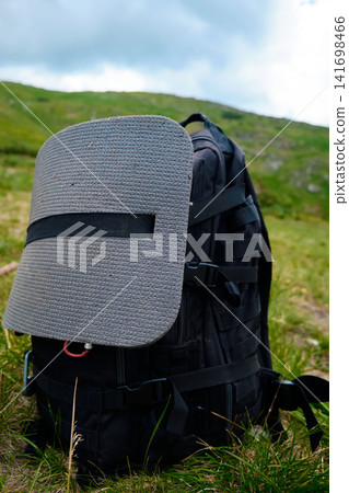 A black tactical tourist backpack with an attached grey foam seat pad stands on the green grass of a mountain hill during a summer trekking trip. A black tactical tourist backpack with an attached grey foam seat pad stands on the green grass of a mountain hill during a summer trekking trip. 141698466