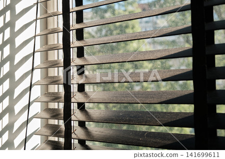 Close-up shot of dark horizontal wooden venetian blinds installed on a window on a sunny day. Sunlight streams through the slats, casting geometric striped shadows on the white wall Close-up shot of dark horizontal wooden venetian blinds installed on a window on a sunny day. Sunlight streams through the slats, casting geometric striped shadows on the white wall 141699611