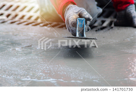 Mason hand in glove smoothing new concrete with a professional trowel tool. Skilled masonry worker performing surface finishing for pavement or sidewalk construction on site. Mason hand in glove smoothing new concrete with a professional trowel tool. Skilled masonry worker performing surface finishing for pavement or sidewalk construction on site. 141701730