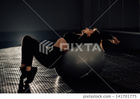 Athletic female performs a back stretch on a stability ball, highlighting balance and wellness. The moody lighting emphasizes her silhouette and the contemporary fitness environment Athletic female performs a back stretch on a stability ball, highlighting balance and wellness. The moody lighting emphasizes her silhouette and the contemporary fitness environment 141701775