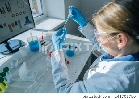 A researcher in a white lab coat uses a pipette to transfer blue solution during a laboratory experiment. Laboratory glassware, test tubes, and digital data analysis are present on the workspace A researcher in a white lab coat uses a pipette to transfer blue solution during a laboratory experiment. Laboratory glassware, test tubes, and digital data analysis are present on the workspace 141701790