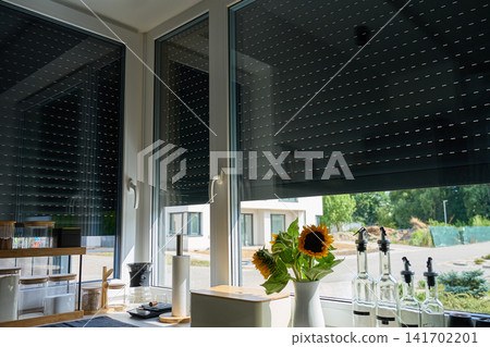 Roller shutters are partly lowered over kitchen windows beside sunflowers, jars and glass bottles Roller shutters are partly lowered over kitchen windows beside sunflowers, jars and glass bottles 141702201