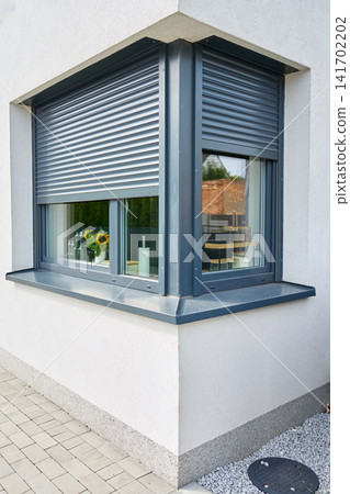 Partially lowered roller shutters cover two corner windows on a modern house, with kitchen items visible inside Partially lowered roller shutters cover two corner windows on a modern house, with kitchen items visible inside 141702202