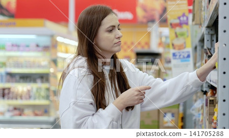 Young woman shopping groceries in supermarket reading product information on shelves, pointing at groceries while making purchasing decisions in retail store Young woman shopping groceries in supermarket reading product information on shelves, pointing at groceries while making purchasing decisions in retail store 141705288