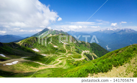 Wide landscape view of a green mountain range in summer, Rosa Khutor resort, Sochi, Russia. Winding dirt road and trails on alpine hills with patches of snow. Blue sky over distant peaks Wide landscape view of a green mountain range in summer, Rosa Khutor resort, Sochi, Russia. Winding dirt road and trails on alpine hills with patches of snow. Blue sky over distant peaks 141705674