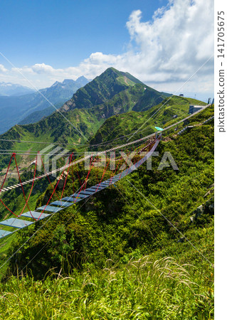 Vertical photo of a glass suspension bridge in the mountains in Sochi, Russia. High altitude rope walkway. Scenic landscape with green peaks at Rosa Khutor resort Vertical photo of a glass suspension bridge in the mountains in Sochi, Russia. High altitude rope walkway. Scenic landscape with green peaks at Rosa Khutor resort 141705675