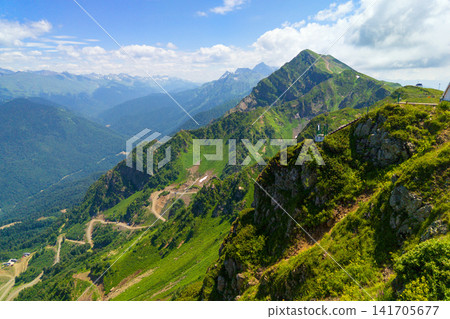 Panoramic view of green mountain peaks under a blue sky in Sochi, Russia. Winding road on a steep grassy slope in a high altitude landscape. Summer travel, nature and adventure Panoramic view of green mountain peaks under a blue sky in Sochi, Russia. Winding road on a steep grassy slope in a high altitude landscape. Summer travel, nature and adventure 141705677