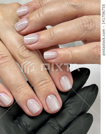 Closeup of a womans hand with short glossy nude nails and glitter gel polish. Professional manicure in beauty salon. Hand in black glove holding client's fingers. Soft pink tone, minimalist and clean Closeup of a womans hand with short glossy nude nails and glitter gel polish. Professional manicure in beauty salon. Hand in black glove holding client's fingers. Soft pink tone, minimalist and clean 141706758