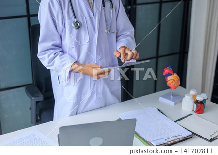 Cardiologist doctor working in office reading patient health records at desk. 141707811