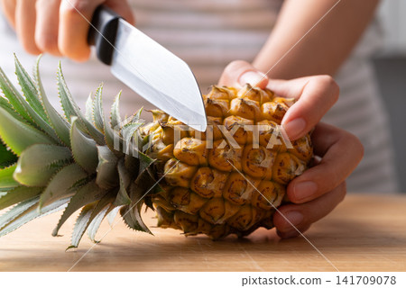 Close-up of a woman's hands cutting a fresh pineapple with a knife on a wooden cutting board Close-up of a woman's hands cutting a fresh pineapple with a knife on a wooden cutting board 141709078
