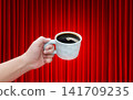 A white ceramic coffee cup is in a man's hand, against a background of a red curtain. 141709235