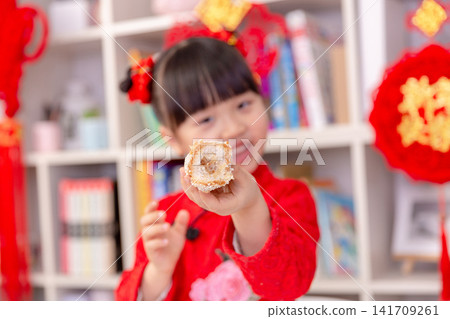 Cute Asian Girl in Traditional Red Dress Eating Chinese New Year Snack,The text on the pendant hanging at the back reads: "Happy New Year." Cute Asian Girl in Traditional Red Dress Eating Chinese New Year Snack,The text on the pendant hanging at the back reads: "Happy New Year." 141709261