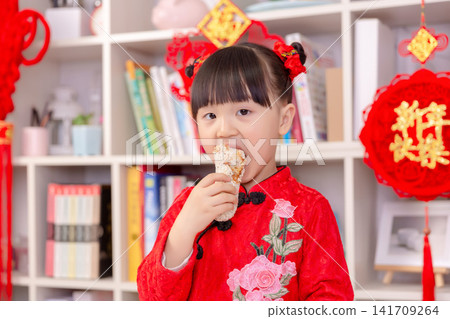 Cute Asian Girl in Traditional Red Dress Eating Chinese New Year Snack,The text on the pendant hanging at the back reads: "Happy New Year." Cute Asian Girl in Traditional Red Dress Eating Chinese New Year Snack,The text on the pendant hanging at the back reads: "Happy New Year." 141709264