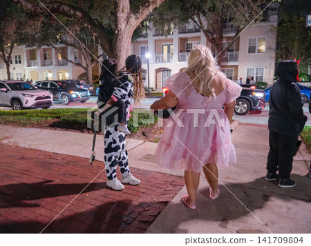 Families wearing Halloween costumes gather outdoors for trick or treating on a neighborhood street at night. Festive celebrations and decorated homes create a lively Halloween atmosphere. Families wearing Halloween costumes gather outdoors for trick or treating on a neighborhood street at night. Festive celebrations and decorated homes create a lively Halloween atmosphere. 141709804
