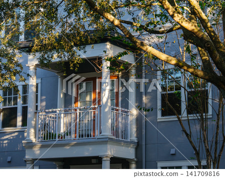 A contemporary apartment building features a balcony framed by leafy trees and warm sunlight. The elegant residential architecture creates a peaceful urban living environment. A contemporary apartment building features a balcony framed by leafy trees and warm sunlight. The elegant residential architecture creates a peaceful urban living environment. 141709816
