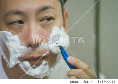一位四十多歲的男子在臉頰上塗抹剃鬚泡沫,然後用T型剃刀進行晨間剃須。 一位四十多歲的男子在臉頰上塗抹剃鬚泡沫,然後用T型剃刀進行晨間剃須。 141709953