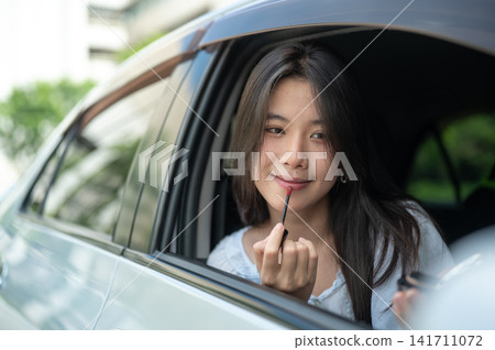 Young Asian woman checking her makeup in mirror from the car window before leaving a home apartment. Young Asian woman checking her makeup in mirror from the car window before leaving a home apartment. 141711072