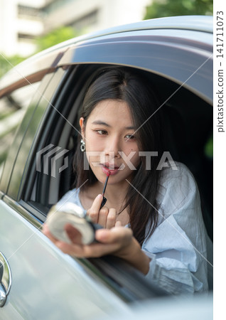 Young Asian woman carefully applying lipstick as she looking into a mirror while sitting in her car. Young Asian woman carefully applying lipstick as she looking into a mirror while sitting in her car. 141711073