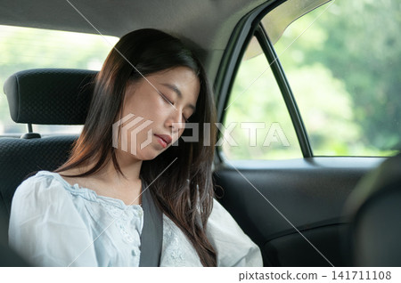 Young Asian woman resting with eyes closed in back seat of a taxi cab car during a relaxing journey. Young Asian woman resting with eyes closed in back seat of a taxi cab car during a relaxing journey. 141711108