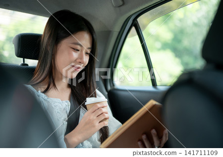 Young Asian woman holding coffee and reading a book while riding in the back seat of a taxi cab car. Young Asian woman holding coffee and reading a book while riding in the back seat of a taxi cab car. 141711109