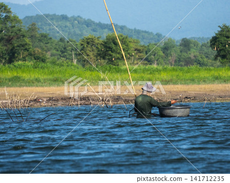 Asian fisherman fishing with a bamboo rod in a calm lake with lush forest and mountains background Asian fisherman fishing with a bamboo rod in a calm lake with lush forest and mountains background 141712235