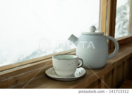 Ceramic pot and cup sit elegantly on wooden ledge with snowy mountains outside Ceramic pot and cup sit elegantly on wooden ledge with snowy mountains outside 141713312