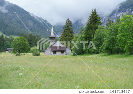Beautiful walking trail in Kandersteg village near Oeschinen Lake. Traditional Swiss wooden chalet, church and summer wildflowers green meadow with stunning Swiss Alps mountains background Beautiful walking trail in Kandersteg village near Oeschinen Lake. Traditional Swiss wooden chalet, church and summer wildflowers green meadow with stunning Swiss Alps mountains background 141713457