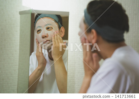 一位四十多歲的男子站在一面三面鏡前,雙手將面膜敷在臉頰上──這是男士的保養流程。 一位四十多歲的男子站在一面三面鏡前,雙手將面膜敷在臉頰上──這是男士的保養流程。 141713661