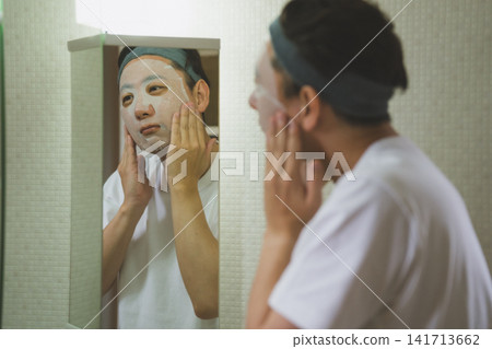 一位 40 歲男子的日常臉部護理程序:在三面鏡前將面膜敷在臉頰上。 一位 40 歲男子的日常臉部護理程序:在三面鏡前將面膜敷在臉頰上。 141713662