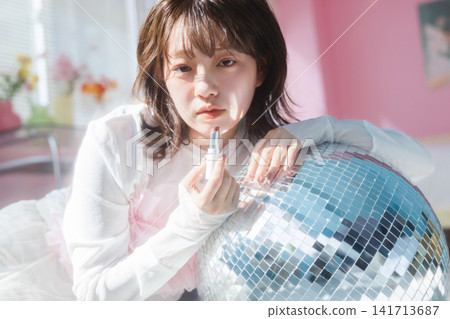A young woman leans against a disco ball, applying lipstick with an ennui expression. A young woman leans against a disco ball, applying lipstick with an ennui expression. 141713687
