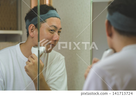 一位 40 歲的男子在三面鏡前使用臉部護理儀護理臉頰：男士護膚。 141713724