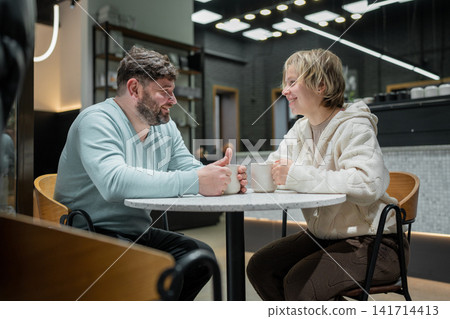 Middle aged man and young woman smiling across marble table in modern coffee shop interior, sharing relaxed daytime conversation with warm industrial lighting, with conversation, smiling, cafe Middle aged man and young woman smiling across marble table in modern coffee shop interior, sharing relaxed daytime conversation with warm industrial lighting, with conversation, smiling, cafe 141714413