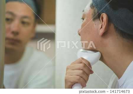 一位 40 歲男子的日常臉部護理程序:在浴室鏡子前使用臉部護理儀按摩下巴。 一位 40 歲男子的日常臉部護理程序:在浴室鏡子前使用臉部護理儀按摩下巴。 141714673