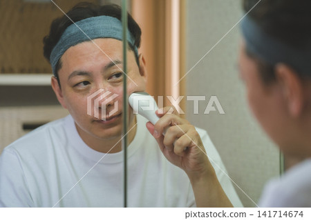 一位40歲的男子對著鏡子,一邊使用臉部美容儀塗抹臉頰。 (男士美容) 一位40歲的男子對著鏡子,一邊使用臉部美容儀塗抹臉頰。 (男士美容) 141714674