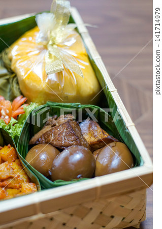 Traditional Indonesian food hamper featuring braised eggs, fried tofu, lontong and festive side dishes arranged in a bamboo basket for Ramadan, Eid, catering and family celebrations. Traditional Indonesian food hamper featuring braised eggs, fried tofu, lontong and festive side dishes arranged in a bamboo basket for Ramadan, Eid, catering and family celebrations. 141714979