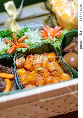 Traditional Indonesian food hamper featuring spicy kentang balado, beef rendang, braised eggs and lontong served in a bamboo basket for Ramadan, Eid, catering and festive family celebrations. Traditional Indonesian food hamper featuring spicy kentang balado, beef rendang, braised eggs and lontong served in a bamboo basket for Ramadan, Eid, catering and festive family celebrations. 141714980