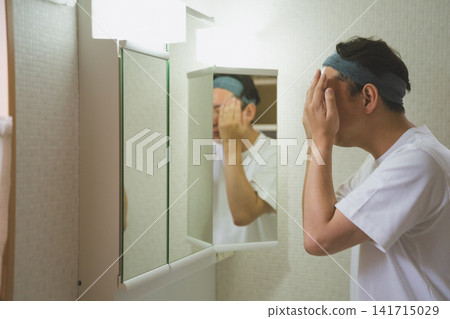 一位四十多歲的男子在三面鏡前用雙手將潤膚露塗抹在臉上,這是他日常臉部護理的一部分。 一位四十多歲的男子在三面鏡前用雙手將潤膚露塗抹在臉上,這是他日常臉部護理的一部分。 141715029